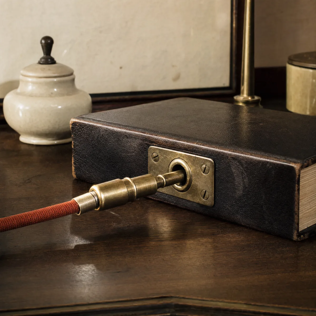 Un enchufe de bronce antiguo, con su cable forrado en tela terracota, conectado a un zócalo metálico incrustado en el lomo de un libro encuadernado en cuero